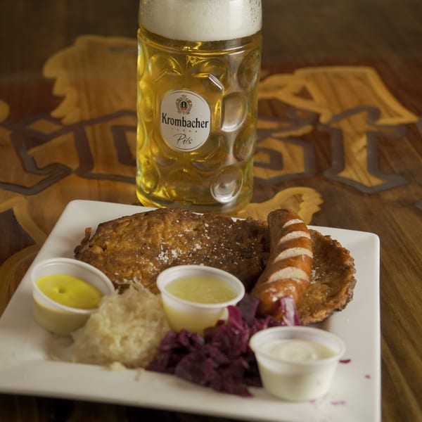 A German platter with schnitzel, sausage, sauerkraut, red cabbage, and sauces, with a Krombacher Pils beer.