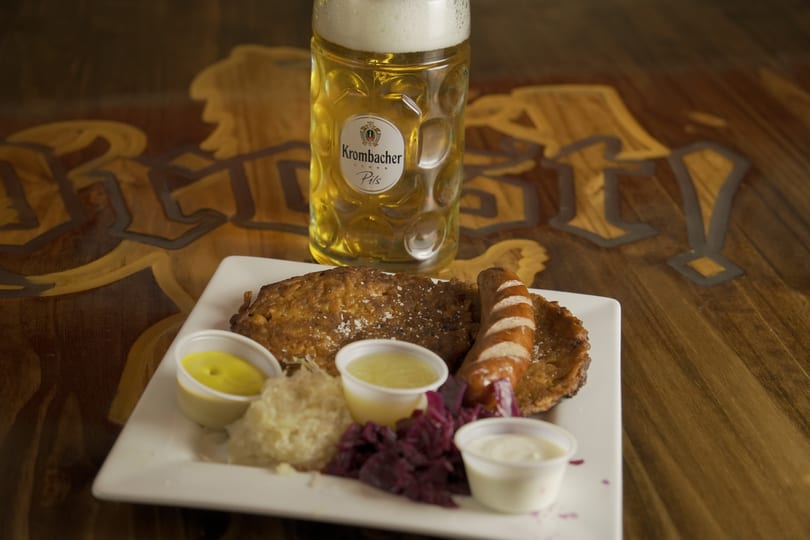 A German platter with schnitzel, sausage, sauerkraut, red cabbage, and sauces, with a Krombacher Pils beer.